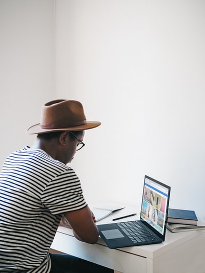 身穿黑白条纹衬衫、头戴棕色帽子的男子坐在椅子上使用微软surface cobalt笔记本电脑